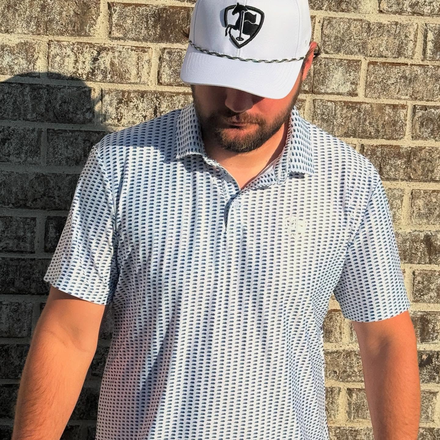 Man wearing a patterned polo shirt and white cap against a brick wall.