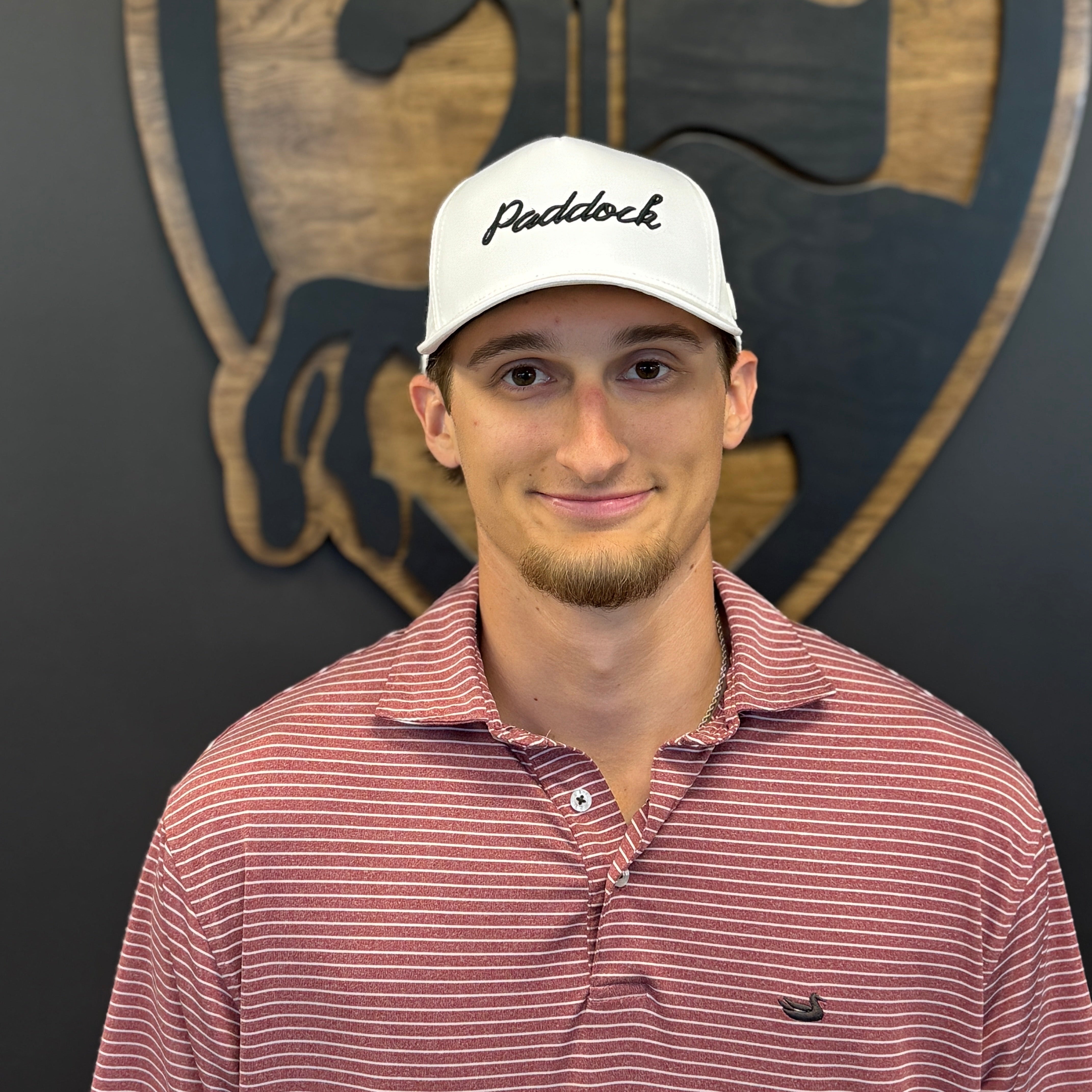 A man wearing a white Paddock brand baseball cap and a red and white striped polo shirt, standing in front of a dark wall with a wooden horse shield logo.